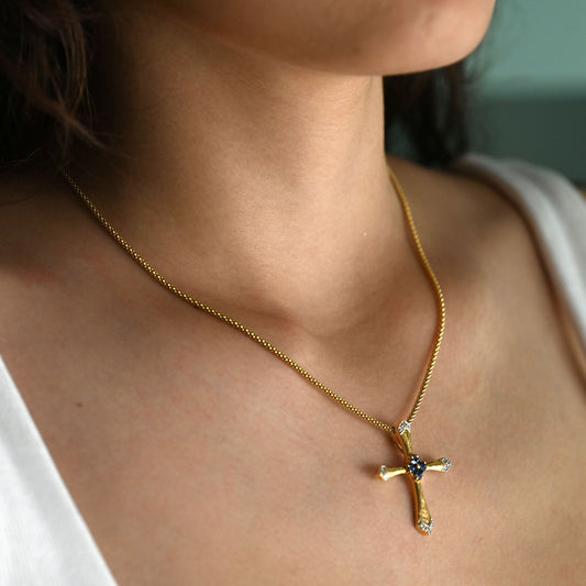 Colgante en forma de cruz latina de oro amarillo de 18kt y combinada con zafiros y brillantes. La cruz destaca por su diseño de líneas curvas estilizadas, un marcado juego de texturas y una cuidada selección de pedreria fina que combina zafiros centrales con pavé de diamantes en los extremos.
