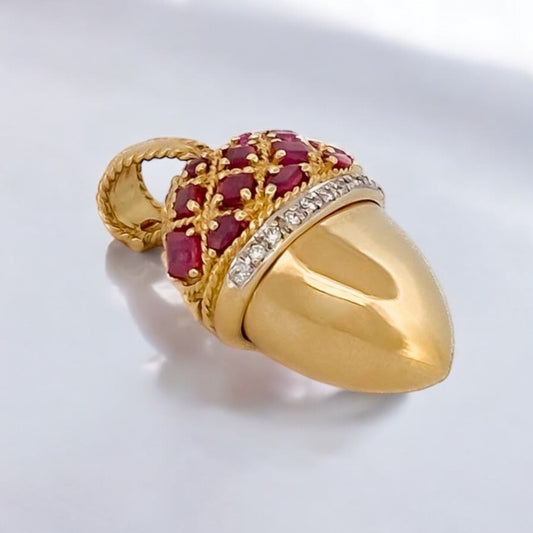 Precioso colgante pastillero de la firma de Joyería Yanes de su colección Malpica, con forma de bellota. Está realizado en oro de 18kt&nbsp; con engaste de rubíes y banda de diamantes. Dispone de una abertura central a modo de pastillero y dispone de una balía de 11,12mm de altura que se puede abrir mediante un cierre de seguridad.